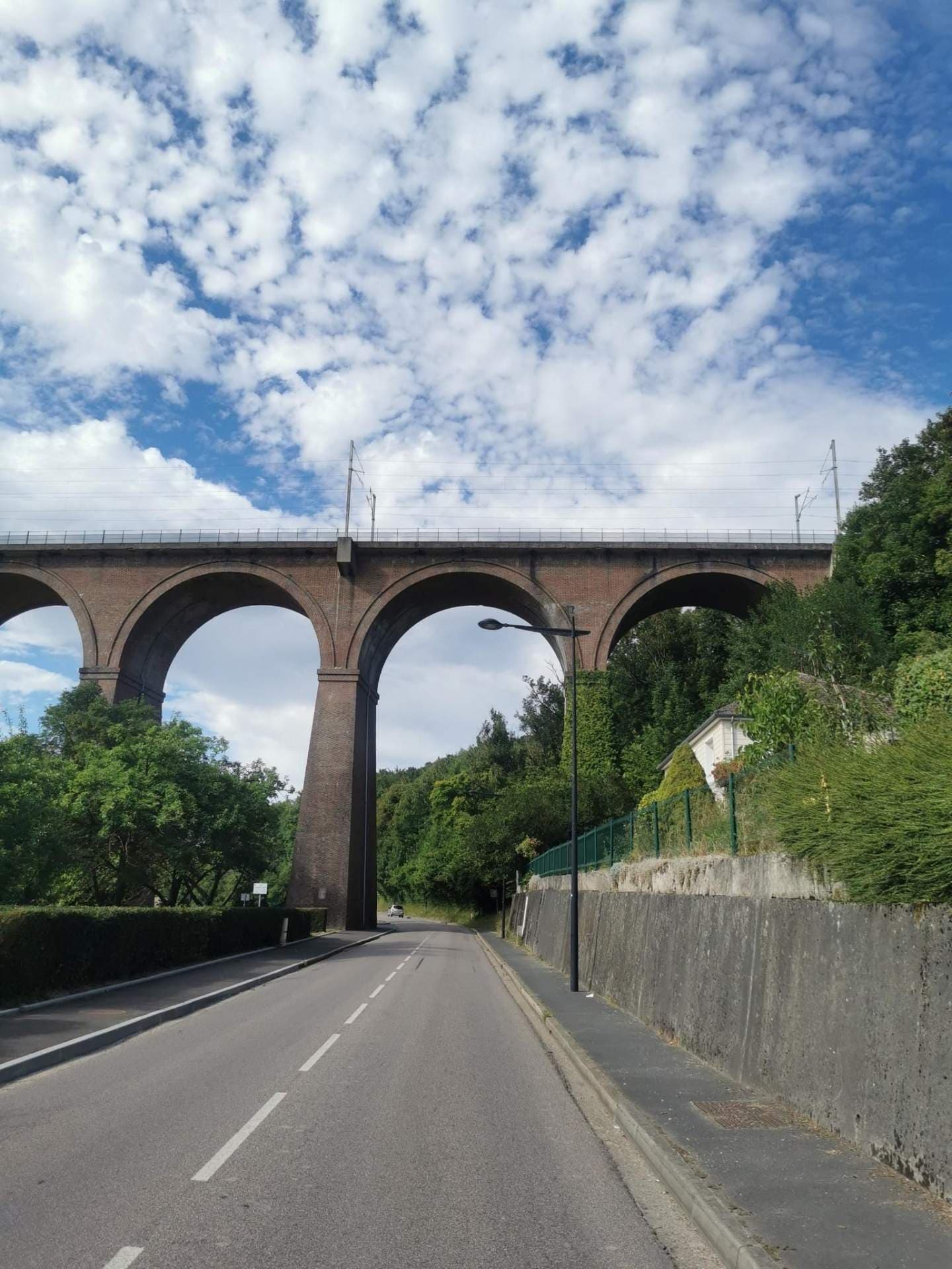 A paved road passes beneath a large brick railway viaduct, bordered by hedges, trees, and a retaining wall under a cloudy sky.
