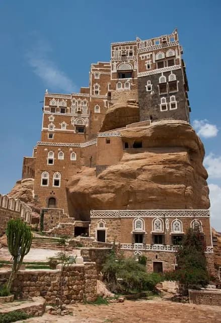A traditional multi-story mud-brick building with white geometric window details built into a rocky hillside under a blue sky.