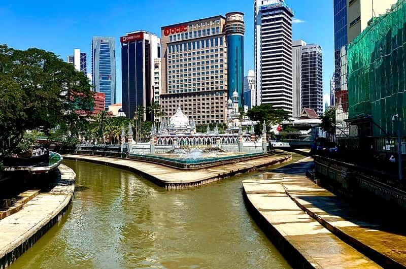 Klang riverfront with a small ornamental pavilion, concrete embankments, and a backdrop of modern office towers under a clear blue sky.