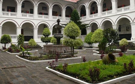 A courtyard featuring a central stone fountain surrounded by manicured gardens and a two-story white building with arched colonnades.