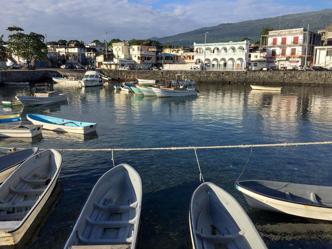 Harbor scene with small boats moored in calm water, buildings and hills in background.