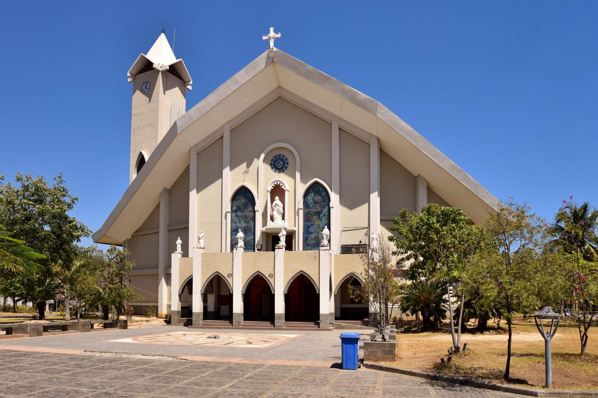 Beige cathedral exterior with steep roof, bell tower, cross, stained glass, and statues, set in a paved plaza with tropical trees under a clear blue sky