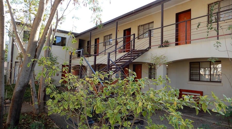 Exterior view of a two-story guesthouse with cream walls, wooden doors, and metal railings, surrounded by trees and greenery.