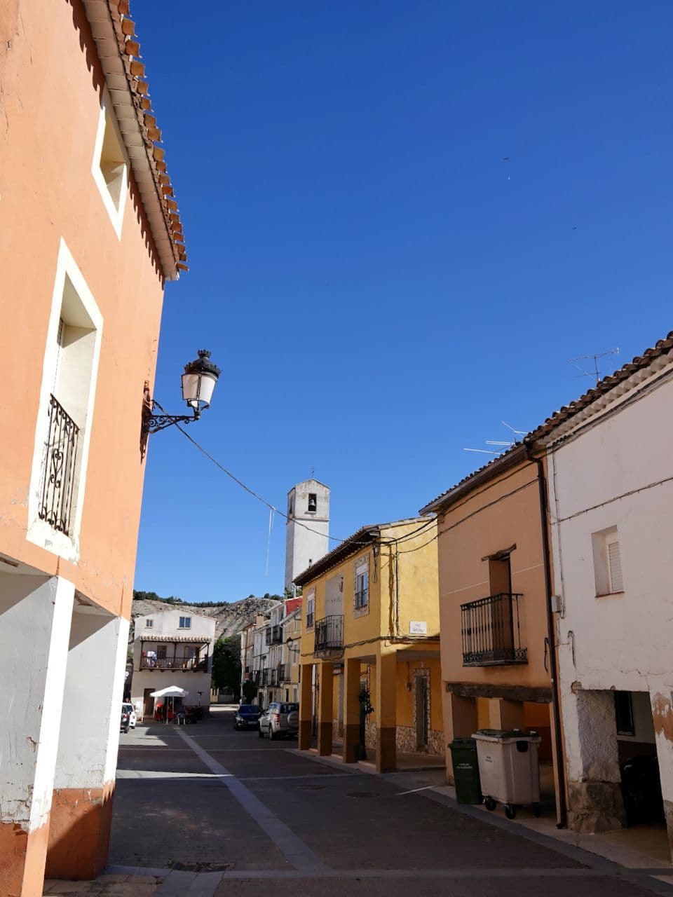 A narrow street in a rural Spanish town with traditional houses, a church tower, and a clear blue sky.