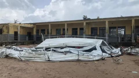 A single-story yellow building with a damaged white tarp structure in the foreground, set against a cloudy sky.