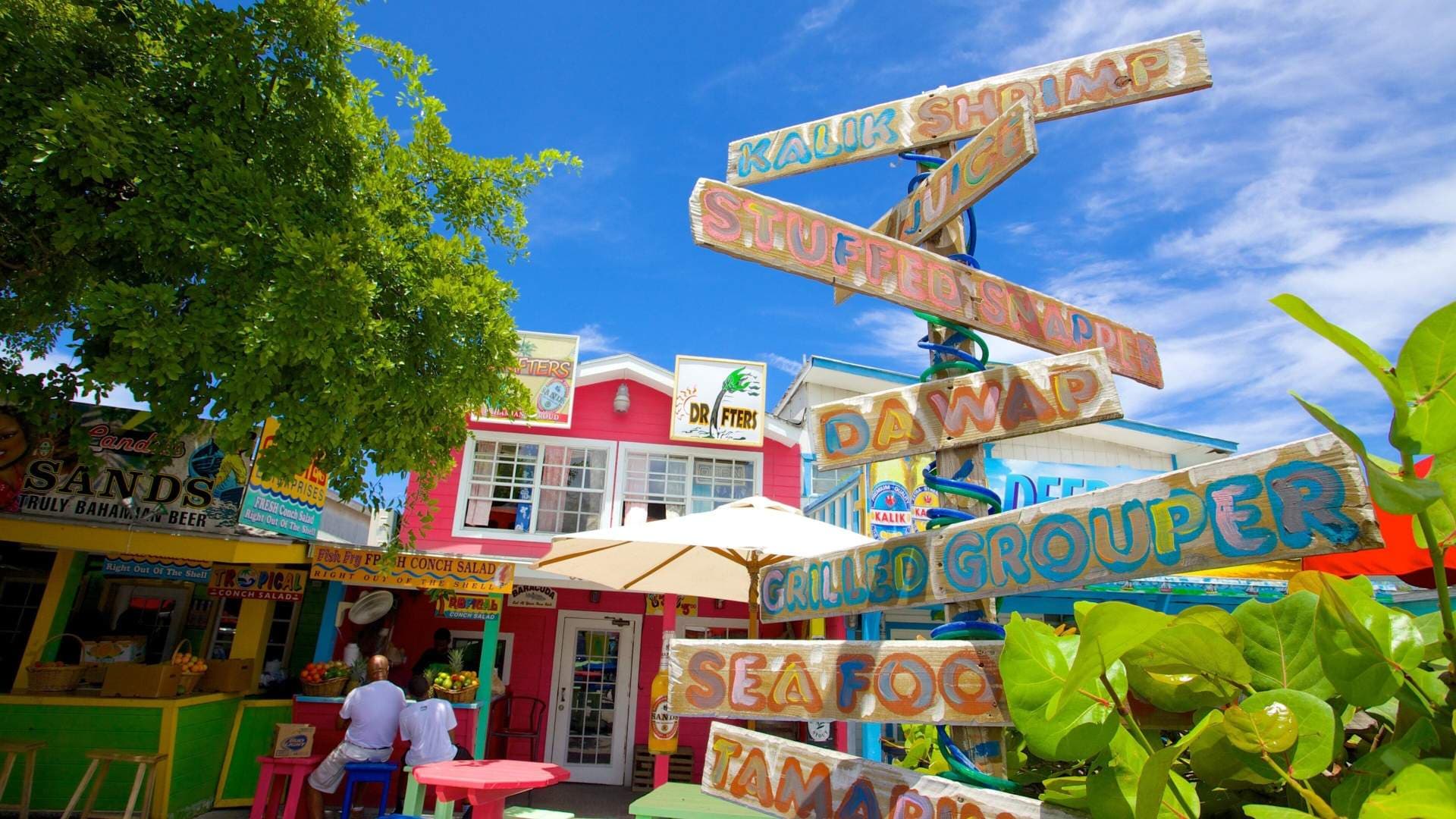Colorful wooden signs advertising seafood and drinks stand in the foreground of a vibrant Bahamian street scene with bright buildings and lush greenery.