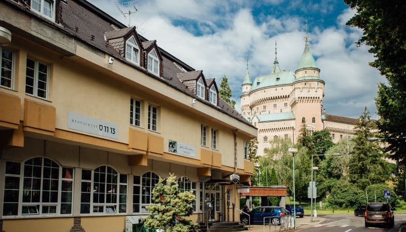 Exterior of a yellow railway station building with a sign, set against a backdrop of a large castle with green spires in the Trenčín Region.