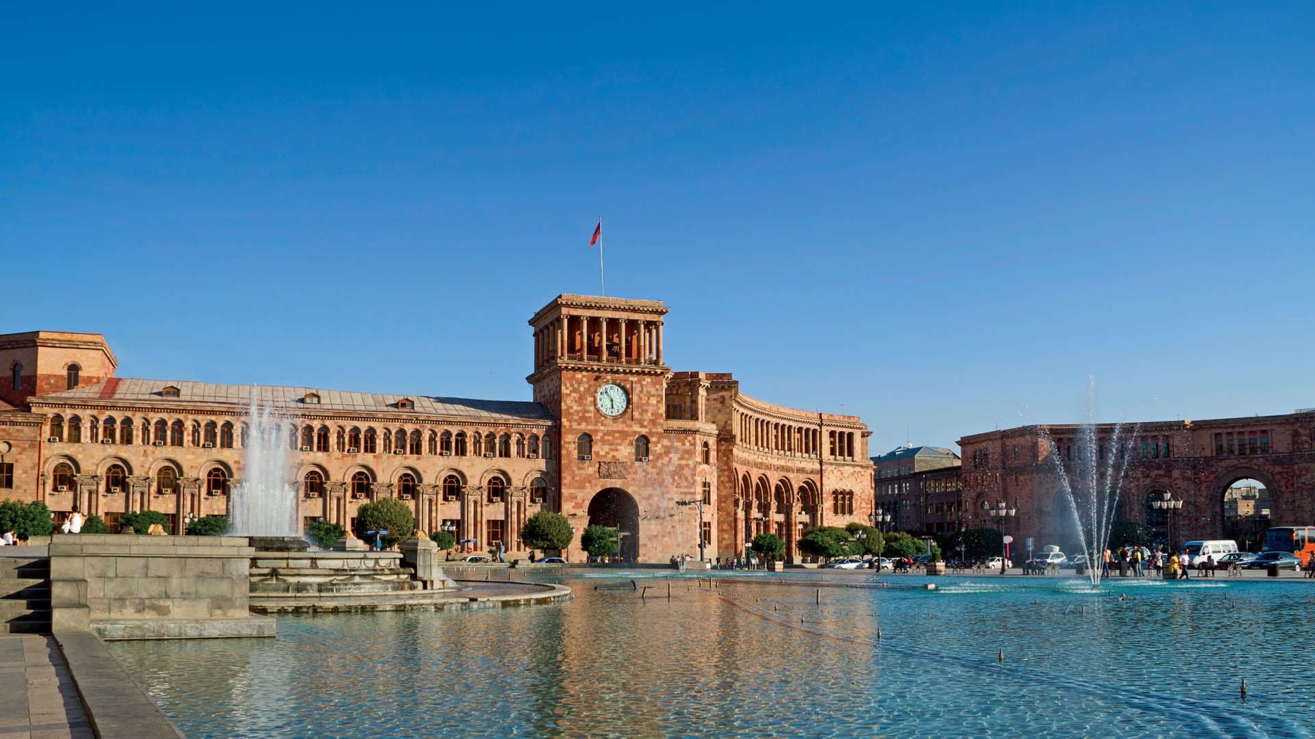 Wide-angle photograph of a large stone civic building with a clock tower and flag, fronted by a plaza fountain under a clear blue sky.