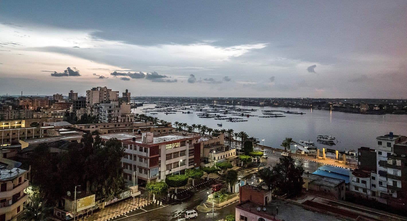 Aerial view of a Nile Delta river landscape at dusk, with urban buildings, palm-lined embankments, boats, and aquaculture pens on the water.