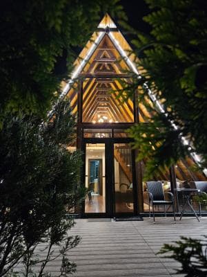 Night view of a modern A-frame wooden guesthouse exterior with illuminated glass facade, patio furniture, and surrounding foliage.