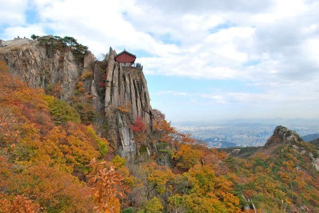 Autumn foliage covers rocky slopes with a small red-roofed pavilion perched on a cliff, overlooking a hazy city skyline.