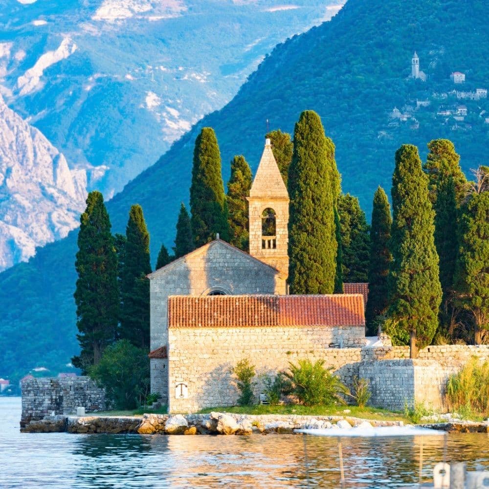 Stone church with bell tower on a small island, surrounded by cypress trees, with the Bay of Kotor and mountains in the background.