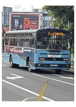 A blue and white double-decker public bus drives on a paved road with white lane markings, featuring a large billboard in the background.