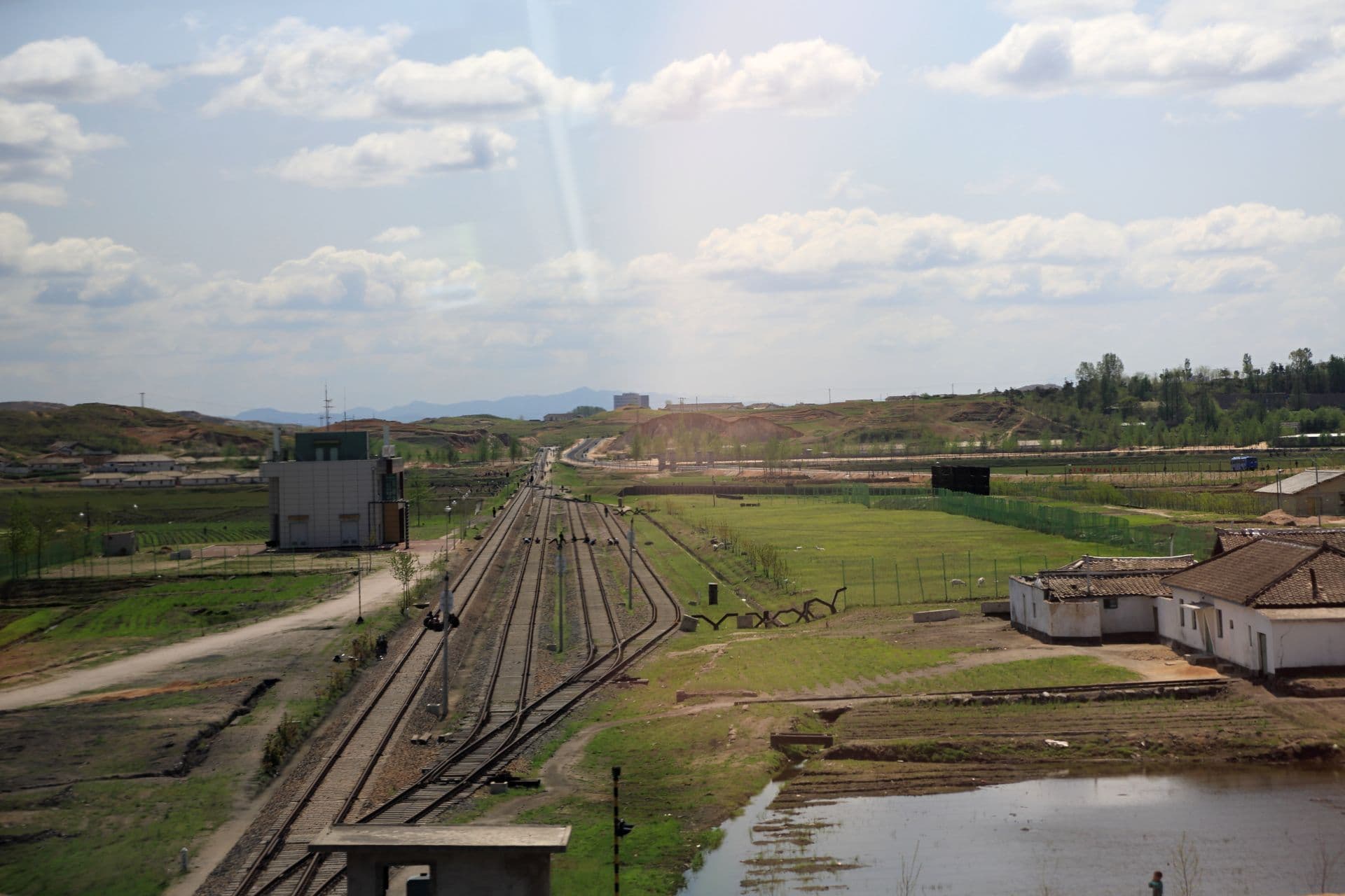 Railway tracks curve through a rural landscape with small buildings, fields, and distant hills under a cloudy sky.