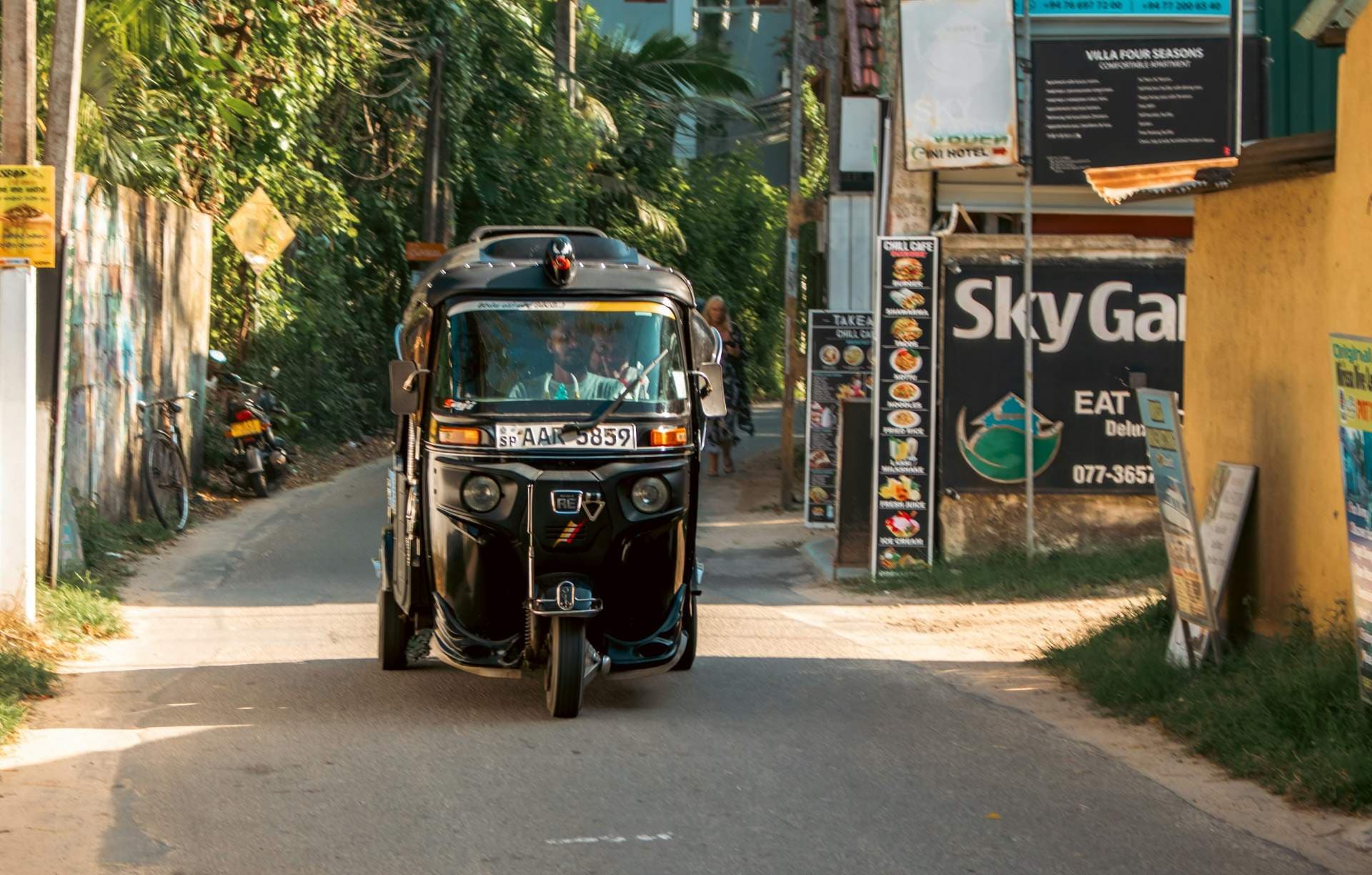 Black tuk-tuk drives down a narrow paved road lined with lush greenery and local business signage.