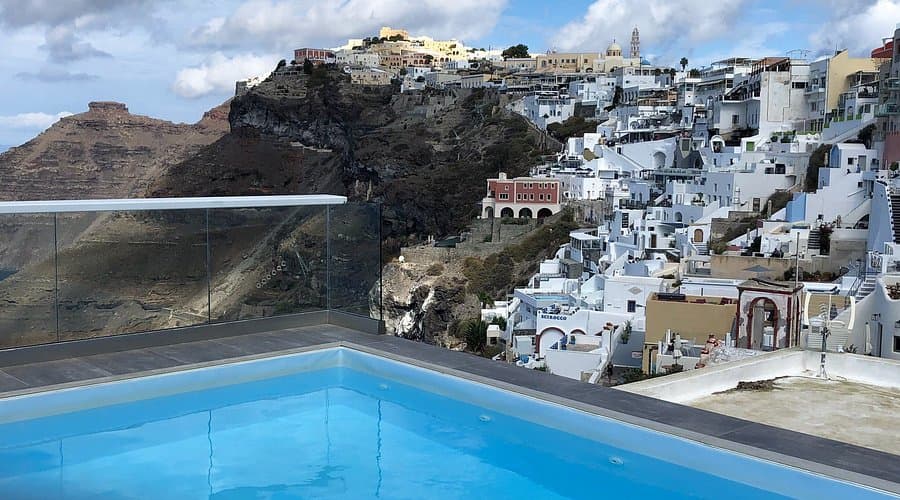 A swimming pool overlooks the whitewashed buildings of Oia village on the Santorini caldera rim, with the volcanic cliff face visible in the background.