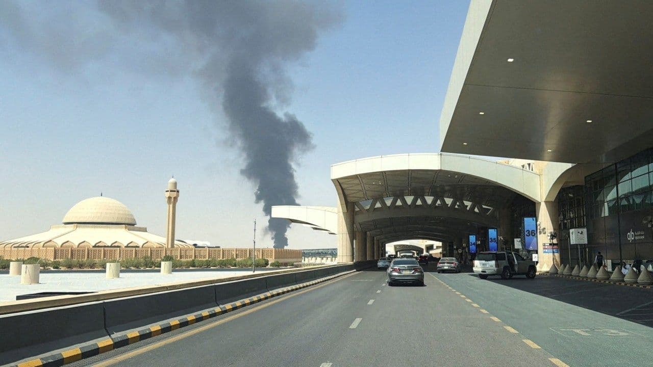 Exterior view of Jizan Regional Airport terminal with cars on the drop-off road and a mosque with dome and minaret to the left