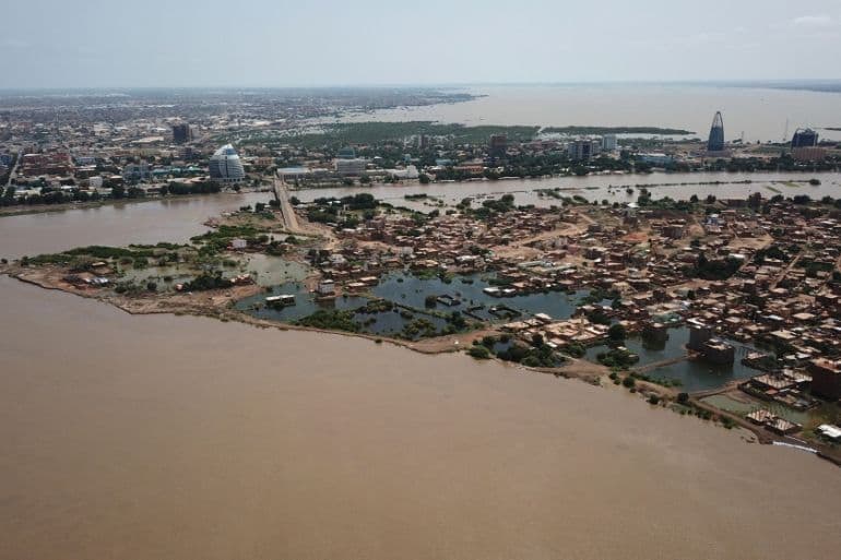 Aerial view of a wide river valley with muddy water, dense settlements, bridges, and a distant city skyline under a hazy sky.