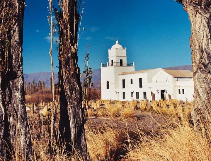 White stone church with bell tower in open pampa landscape, framed by trees and dry grass under blue sky.