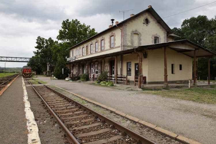 Exterior view of a rural railway station building with tracks and a red train approaching on the left under cloudy skies.