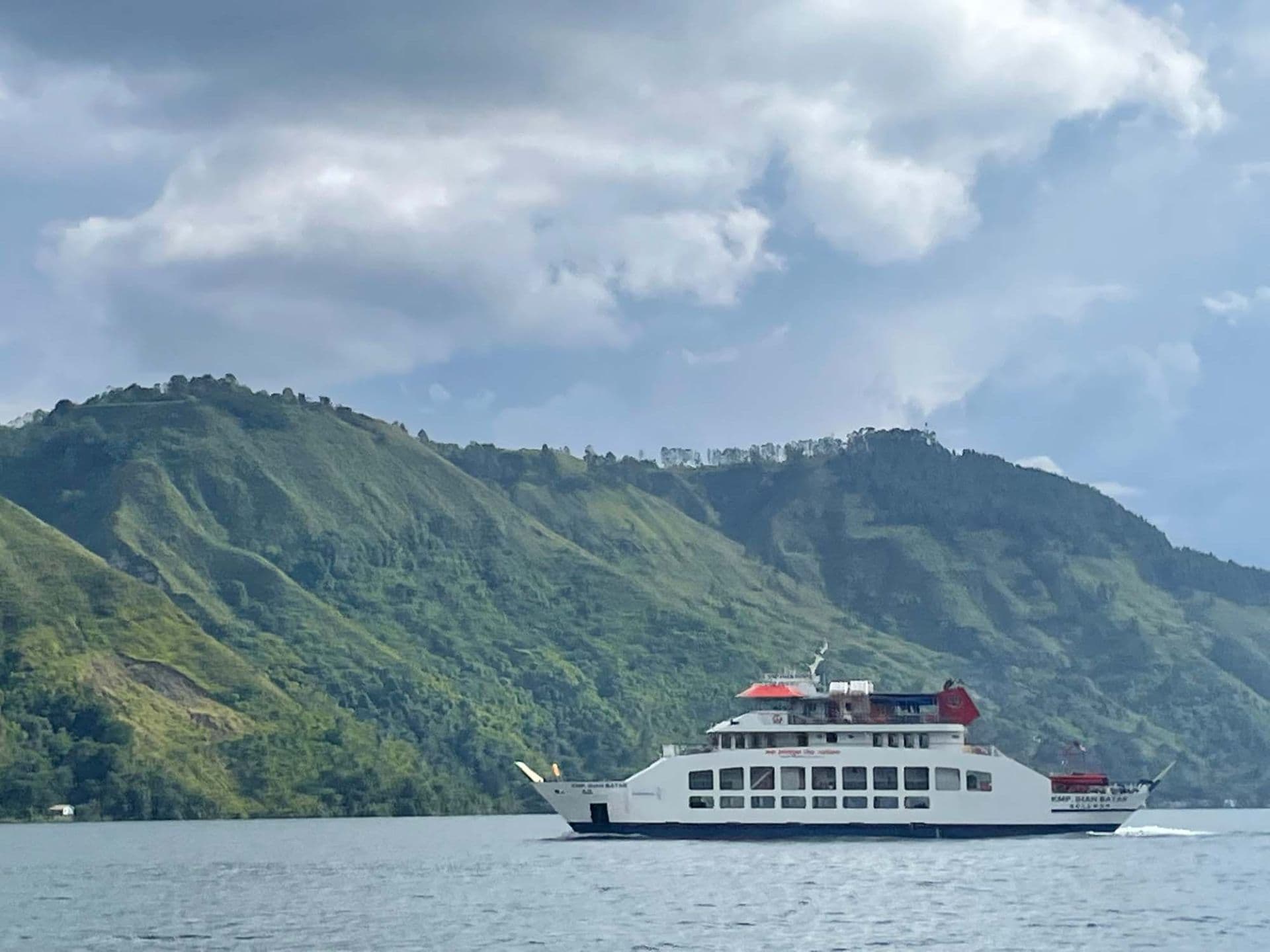 A white passenger ferry navigates calm waters with lush green mountains rising in the background under a cloudy sky.