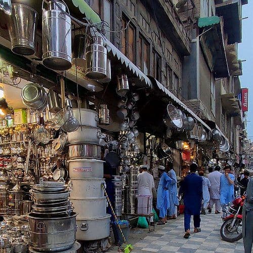 A bustling market street with hanging metal cookware, stacked pots, and pedestrians in traditional blue attire walking past shops.