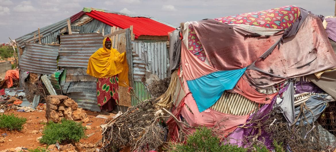 A woman in yellow stands before makeshift corrugated metal and tarp shelters in a rural Somali settlement.