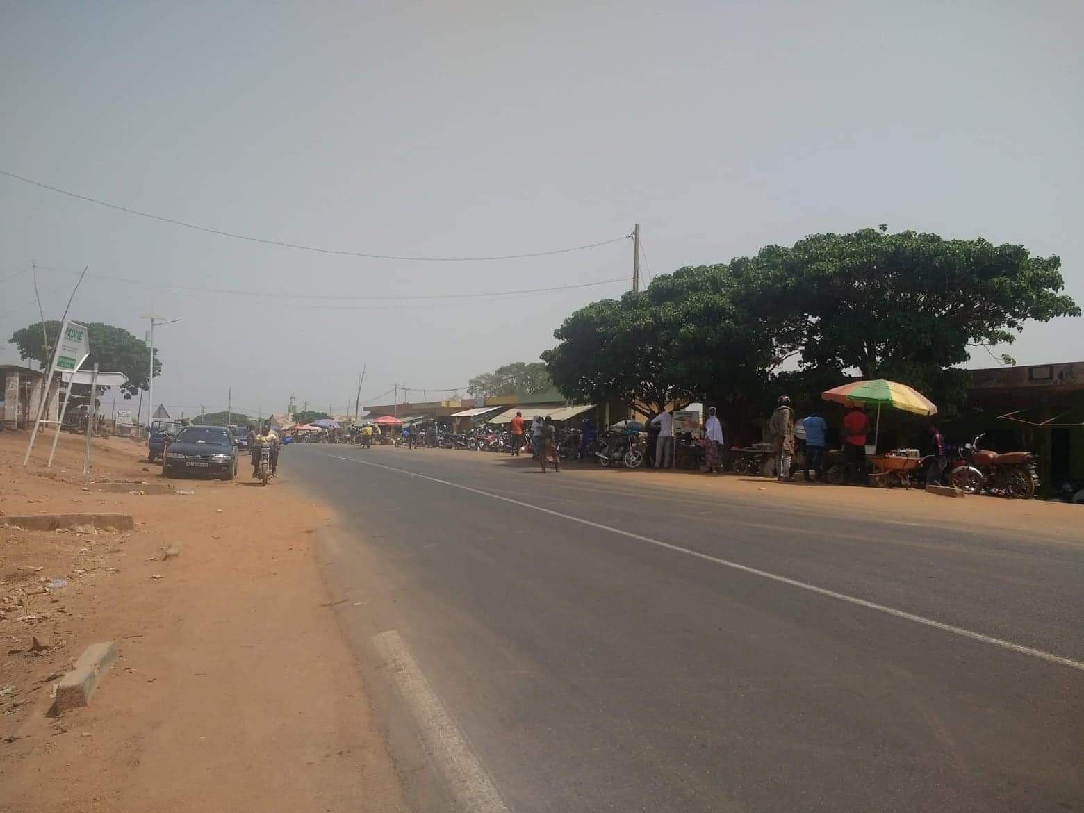 Wide street scene with roadside market stalls, parked motorcycles, a large tree, and a hazy sky.