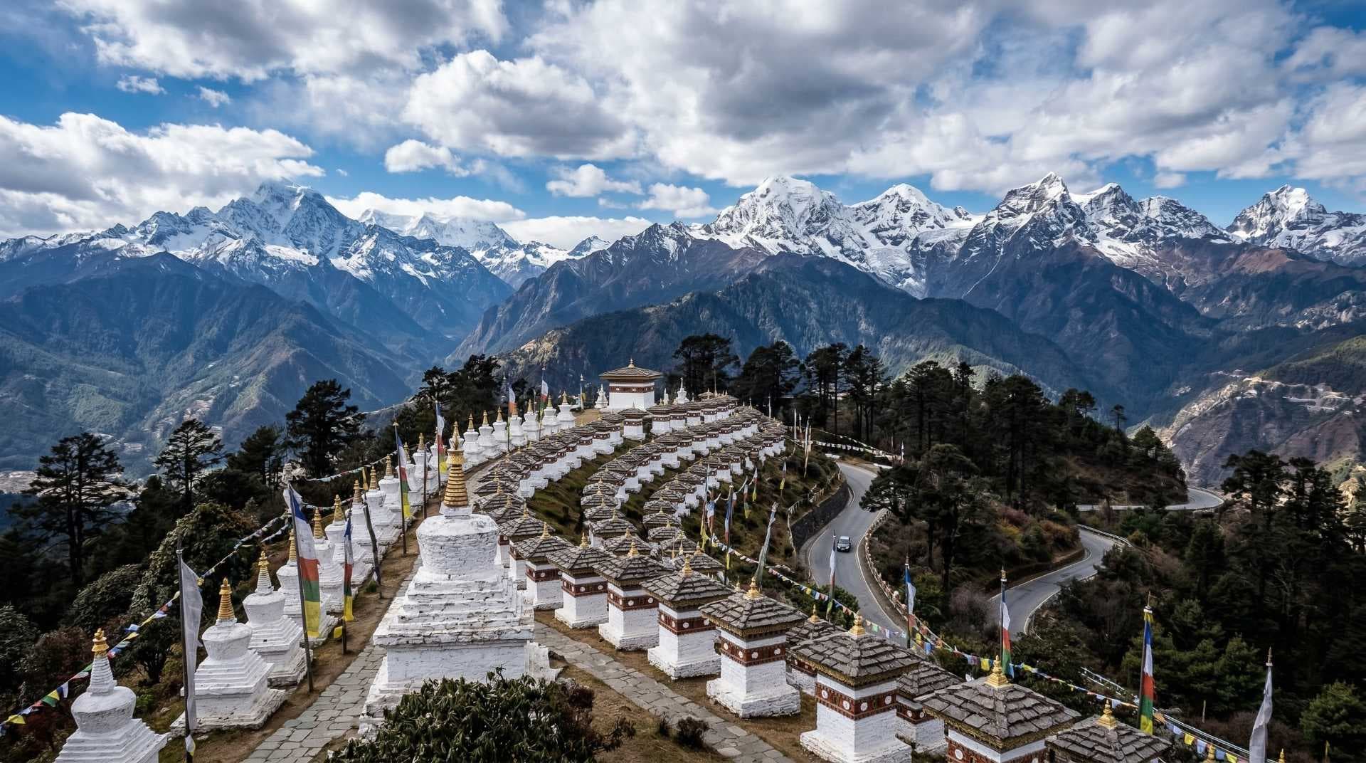 Rows of white chorten stupas with prayer flags line a hillside at Dochula Pass, backed by snow-capped Himalayan peaks and a winding road.