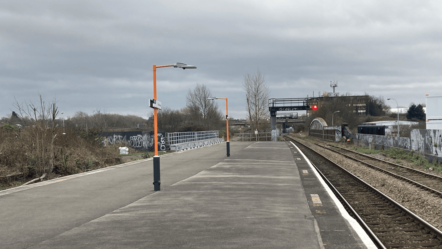 Empty concrete platforms and tracks at Polesworth railway station under an overcast sky, featuring orange lamp posts and graffiti-covered walls.