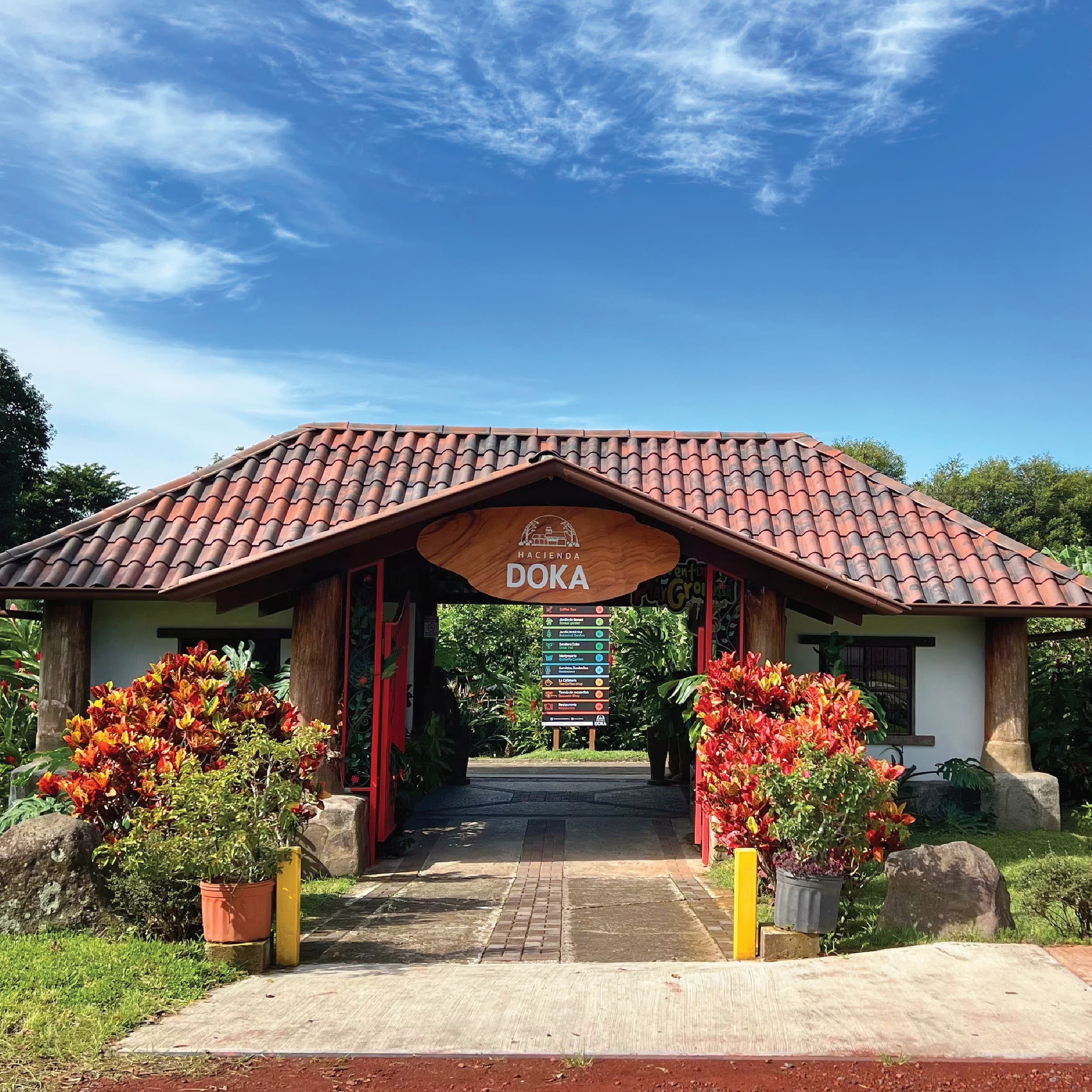 Exterior entrance gate of Hacienda Doka featuring a tiled roof, wooden sign, and vibrant red croton plants under a blue sky.