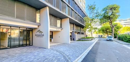 Street view of a modern apartment building with a paved driveway and a parked car, set against a backdrop of trees and blue sky.