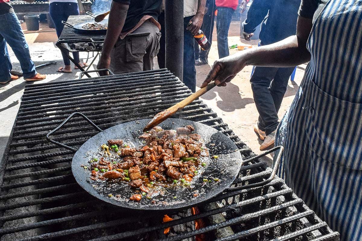 Outdoor charcoal grill with a flat pan of cooked meat and vegetables, people cooking and gathering around the fire.