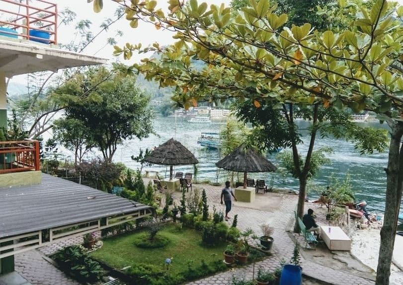 A scenic view of a lakeside resort patio with thatched umbrellas and manicured gardens overlooking Lake Toba, with boats visible on the water.
