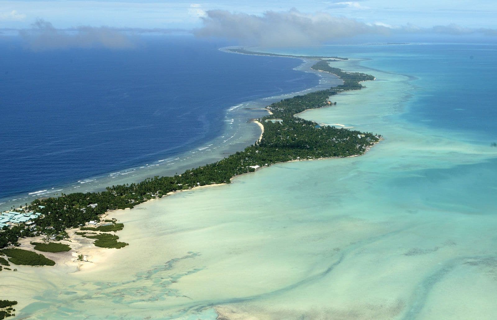 Aerial wide view of a narrow tropical island chain with turquoise lagoon, deep blue ocean, and scattered coastal buildings.