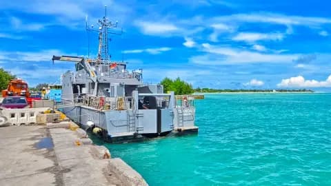 A gray harbor boat moored at a concrete pier beside turquoise water, with vehicles and a small island visible in the background.