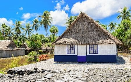 Beachfront scene with thatched-roof village houses, palm trees, and a rocky shoreline under a blue sky.