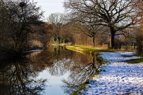 A serene winter landscape featuring a calm river reflecting bare trees, with snow dusting the grassy banks and a path winding through the scene.