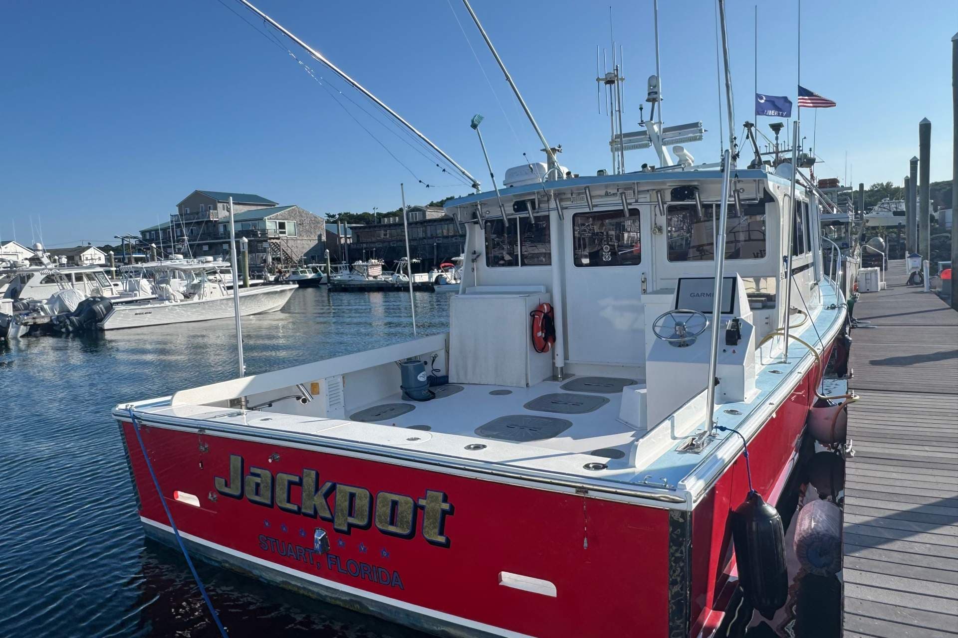 Red fishing boat named Jackpot docked at a working port, with other boats and waterfront buildings in the background.