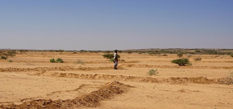 A lone figure stands in a vast, arid landscape with eroded earth and sparse vegetation under a clear blue sky, depicting the terrain of the Bakool region.