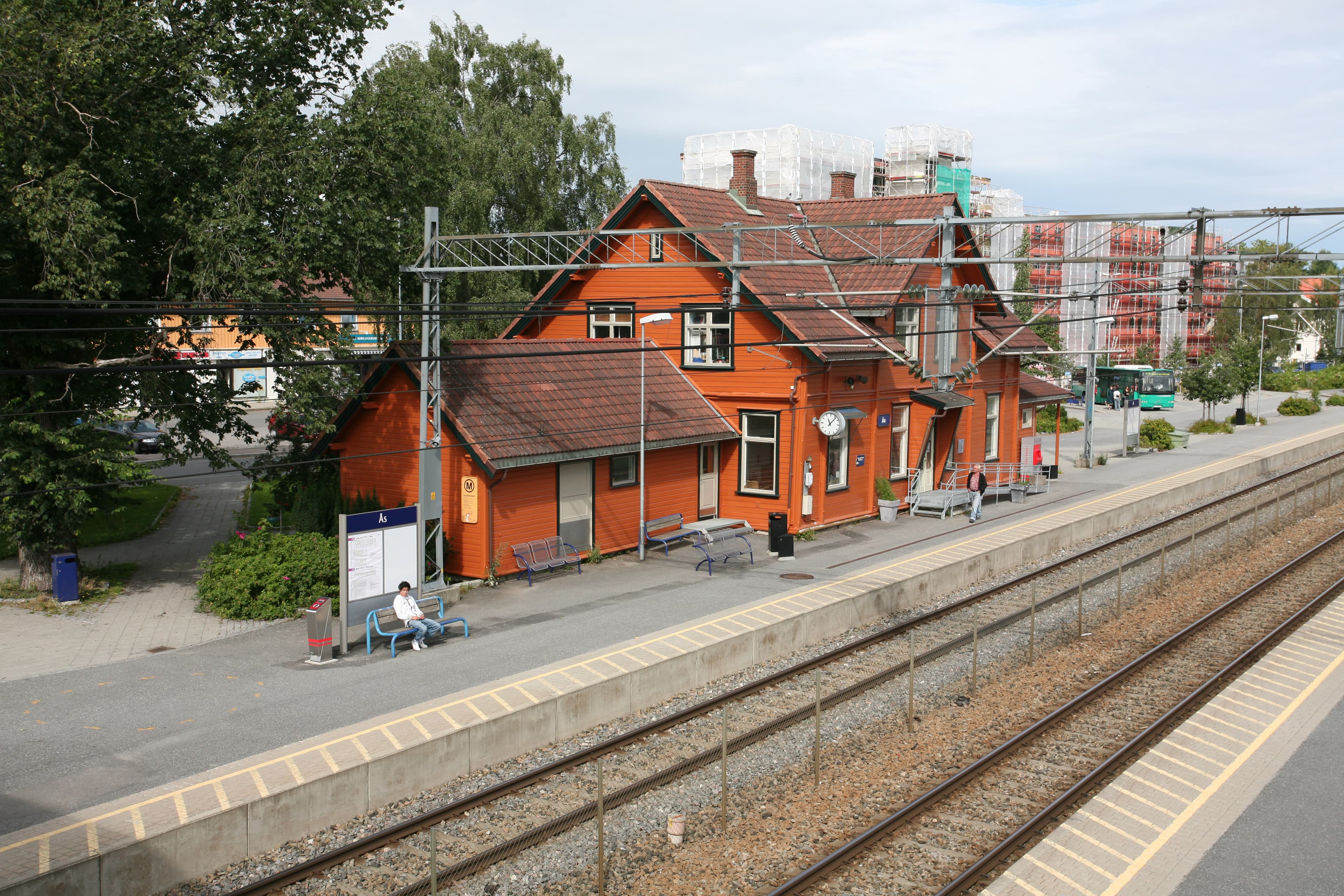 Aerial view of a red wooden train station building with tracks and a platform, featuring a few waiting passengers and overhead wires at Ås Station near Drøbak.