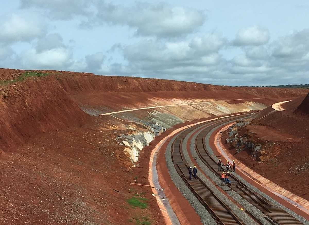 Curving railway tracks cut through a vast red earth landscape in Guinea, with workers visible on the rails under a cloudy sky.