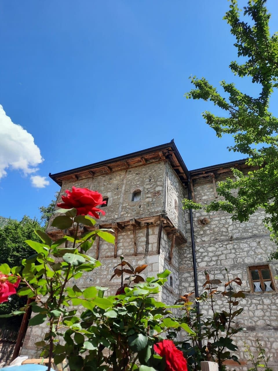 Stone kulla tower with wooden balconies and village houses under a blue sky, framed by red roses and green foliage.