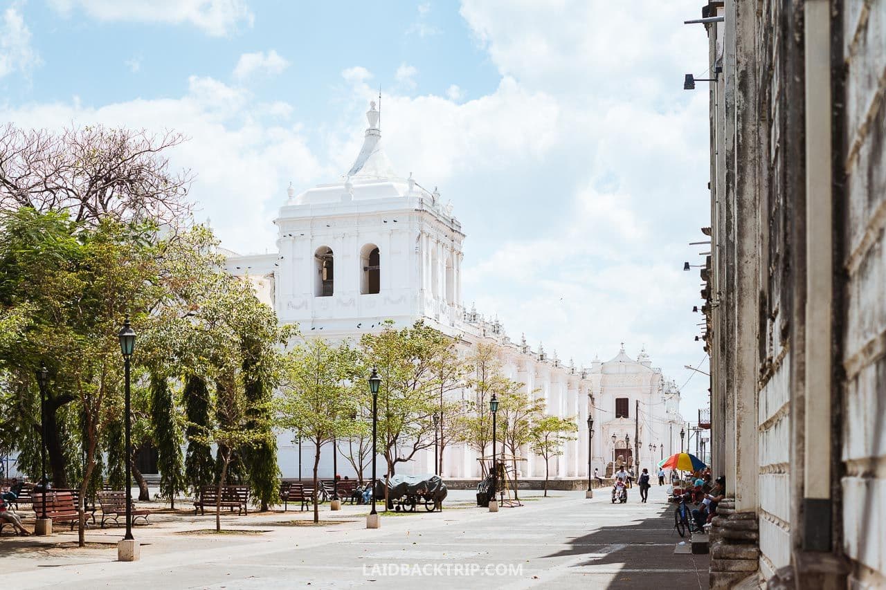 Wide shot of a white colonial church and plaza in Telica, Nicaragua, featuring trees, benches, and distant pedestrians under a blue sky.