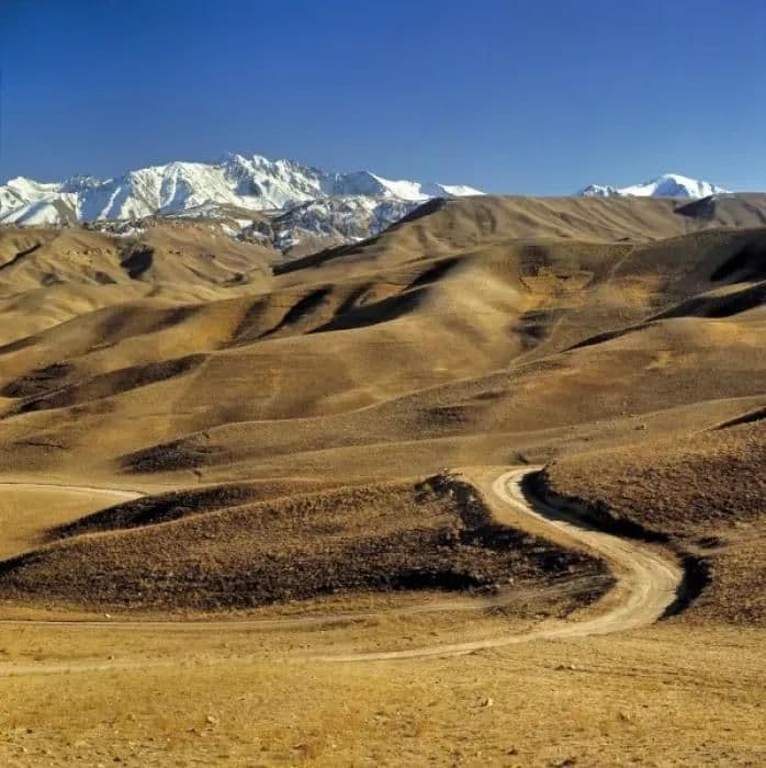 A winding dirt road curves through arid brown foothills toward distant snow-capped mountains under a clear blue sky.