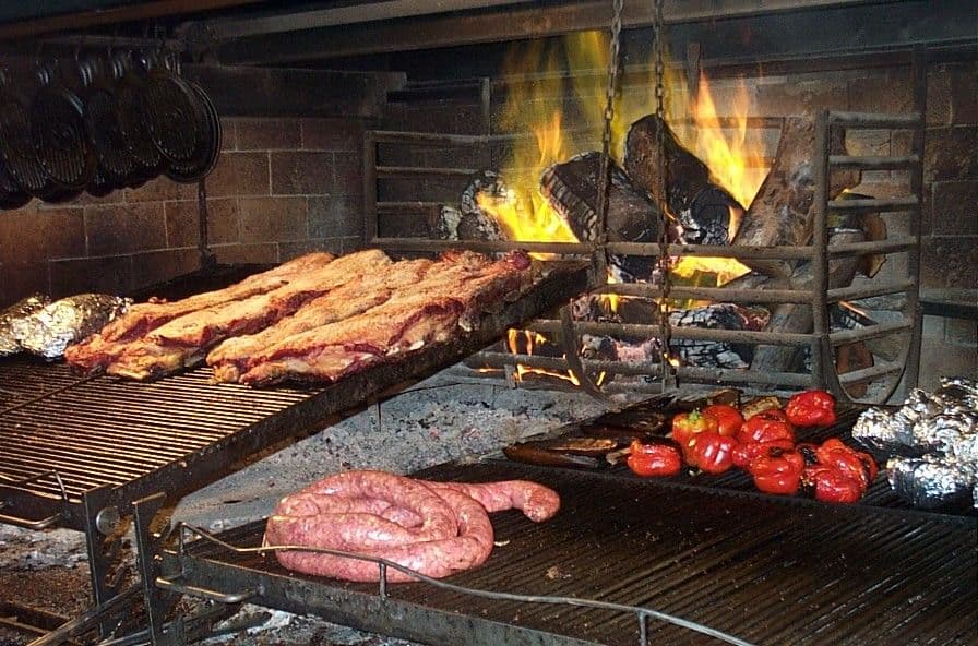 Rural open-fire asado grill with ribs, coiled sausage, and red peppers cooking over wood coals.