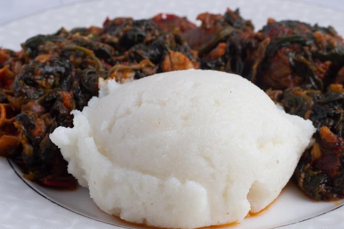 Close-up photograph of a white mound of sadza (maize meal) served with a dark vegetable and meat stew on a white plate.