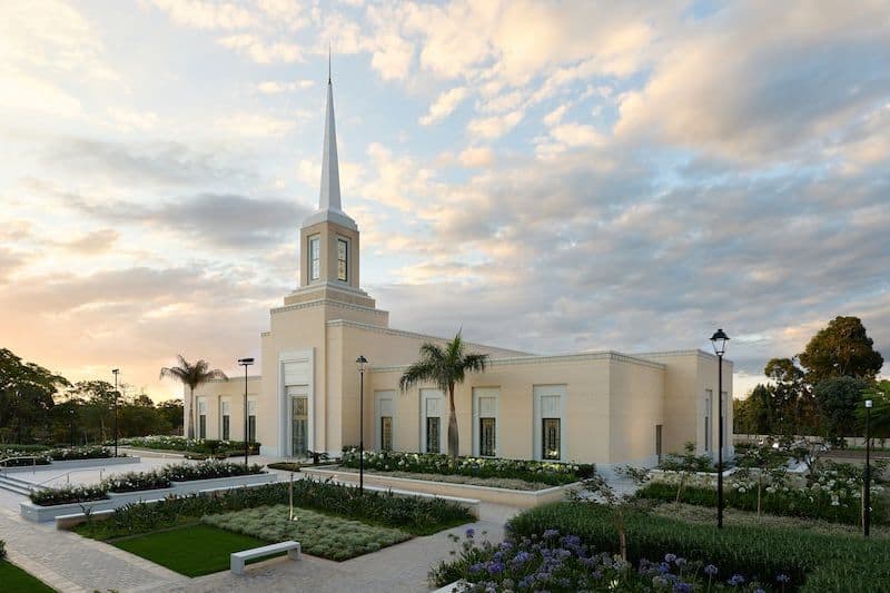 Modest cream-colored church exterior near Marondera District, Zimbabwe with a tall white steeple, surrounded by manicured gardens and palm trees under a cloudy
