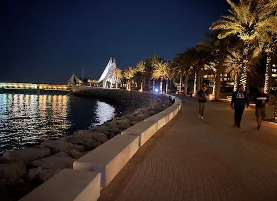 Night view of the Kuwait waterfront promenade near Ash Shāmīyah with palm trees, stone seawall, and an illuminated walkway