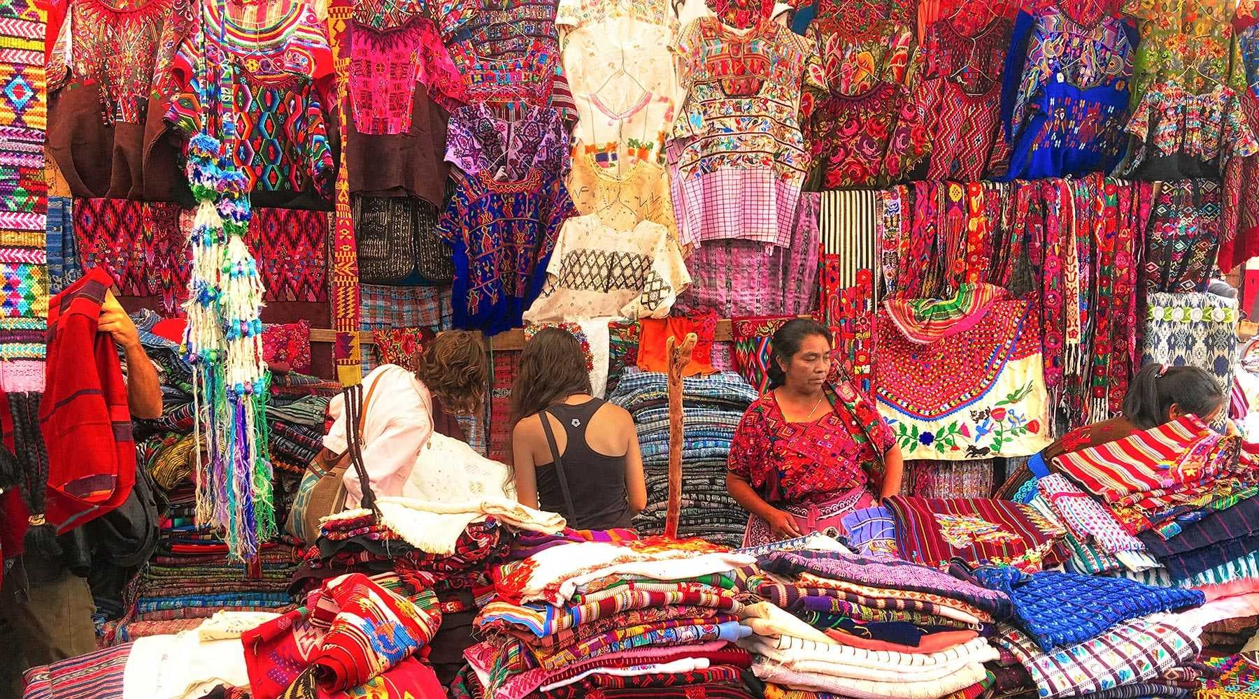 Vibrant market stall displaying colorful woven textiles, embroidered garments, and artisan crafts with vendors and shoppers present.